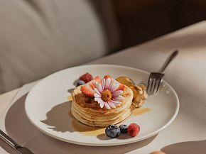 Ein Teller mit Pfannkuchen, garniert mit Beeren und einer Blume, liegt auf einem Tisch. Sonnenlicht fällt sanft auf die Anrichte.