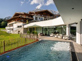 Modernes Hotel mit alpinem Design, eingebettet in grüne Natur, mit Pool im Vordergrund und Bergkulisse im Hintergrund. Sonnige Terrasse mit Sitzgelegenheiten.