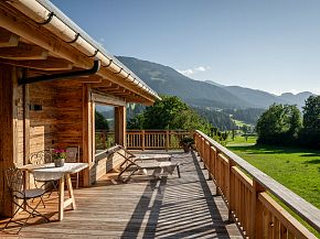 Holzveranda mit Blick auf grüne Wiesen und bewaldete Berge unter blauem Himmel, ausgestattet mit Liegestühlen und einem kleinen Tisch mit Stühlen.