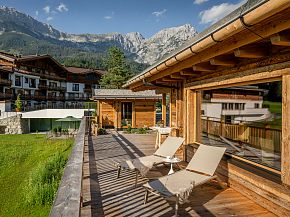 Panorama eines alpinen Hotels mit Holzterrassen, Liegestühlen und Bergblick im Hintergrund. Sonniges Wetter und grüne Umgebung vermitteln Entspannung.
