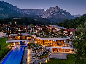Ein Hotel in alpiner Landschaft bei Dämmerung, umgeben von Bergen. Ein beleuchteter Pool ergänzt die gemütliche Atmosphäre des Gebäudekomplexes.