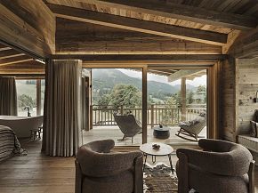 Gemütliches Hotelzimmer mit Holzausstattung und großem Fenster mit Bergblick. Sitzbereich und Balkon laden zum Entspannen ein.