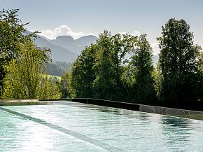 Ein Naturpool im Freien, umgeben von grünen Bäumen und mit Blick auf die sanften Berge im Hintergrund, unter einem klaren blauen Himmel.