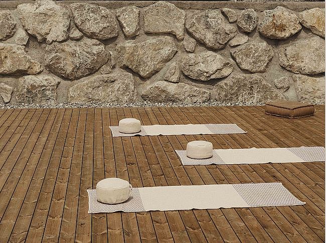 Holzboden mit drei Yoga-Matten und Meditationskissen vor einer Steinmauer. Ruhige und entspannte Atmosphäre für Outdoor-Yoga-Sitzungen.