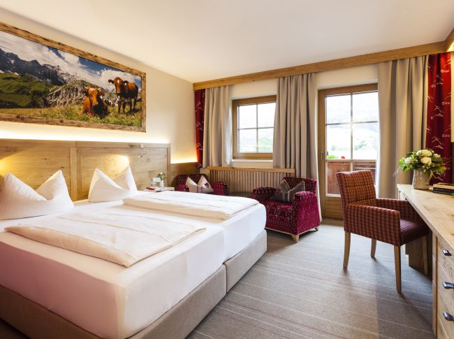 Gemütliches Hotelzimmer mit Doppelbett, rustikalen Holzmöbeln, einem großen Landschaftsfoto an der Wand und einem Balkon mit Blick auf die Berge.