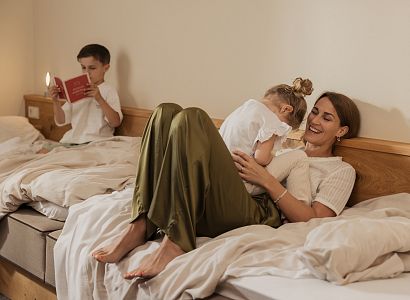 Eine Mutter entspannt mit ihren zwei Kindern in einem gemütlichen Hotelzimmer. Ein Kind liest, während das andere mit der Mutter kuschelt.