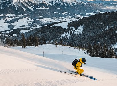 Skifahrer in gelbem Anzug fährt bei Sonnenuntergang eine verschneite Piste hinunter, umgeben von beeindruckender Berglandschaft.